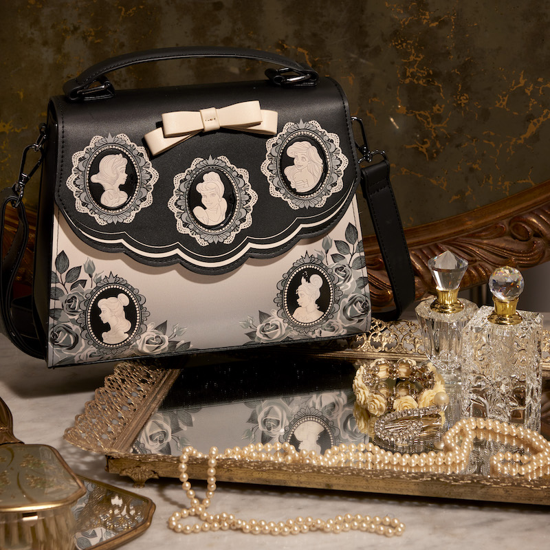 Black and white Loungefly Disney Princess Cameo Porcelain Portraits Crossbody Bag sitting on an try with a pearl necklace and perfume bottles 